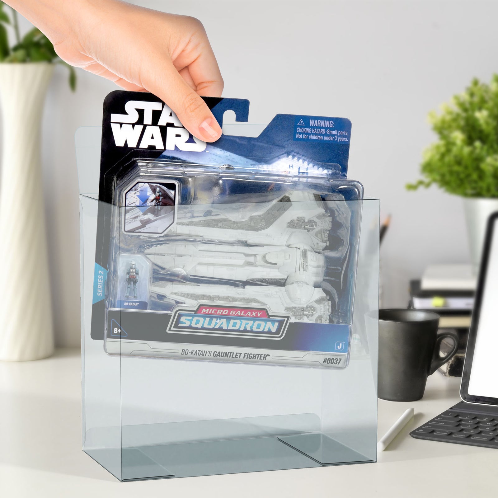 Hand placing Star Wars Micro Galaxy Squadron into PET protector on desk, with laptop and plants in background