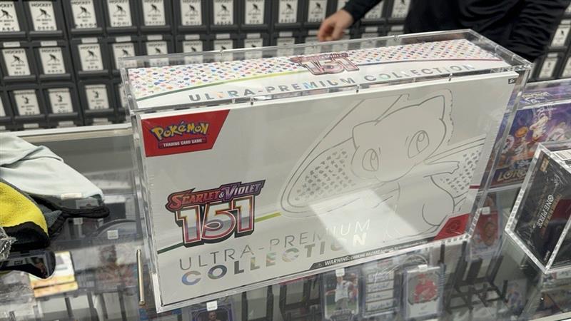 Acrylic case with Pokémon 151 Ultra-Premium Box on a shelf with other items.
