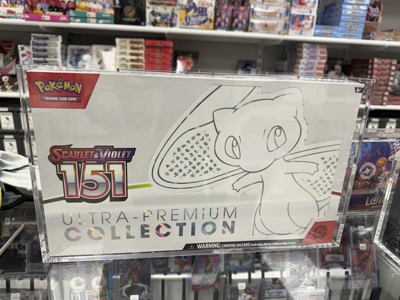 Acrylic display case with a Pokémon 151 Scarlet & Violet Ultra-Premium Collection Box inside, placed on a shelf with various other items.