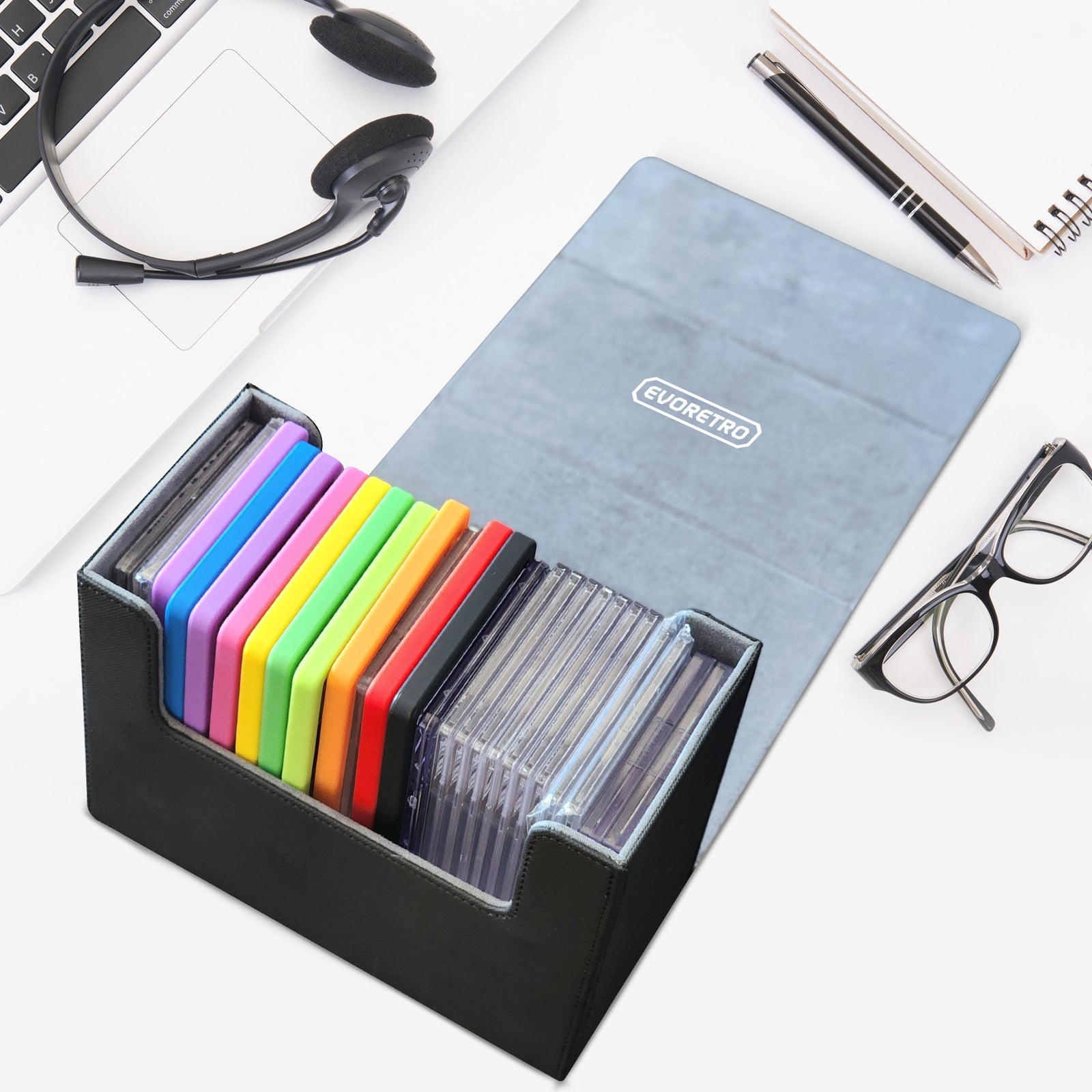 Open graded card holder with colorful slabs on a white desk setup beside laptop, headset, glasses, and notepad